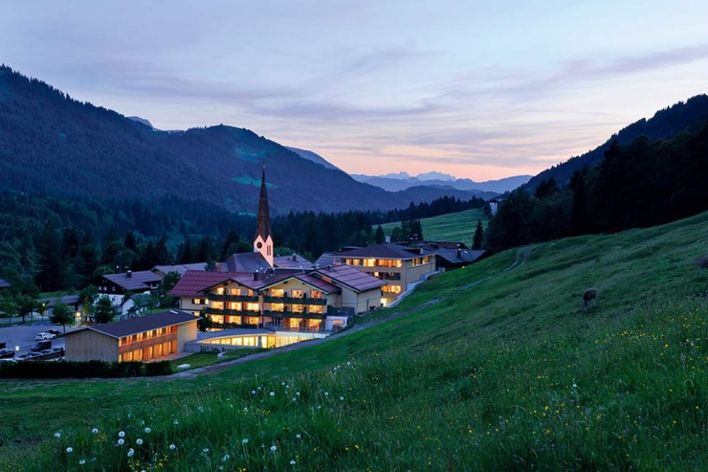 Abendstimmung mit Blick von der Almwiese auf Balderschwang im Allgäu. Vor der Dorfkirche liegt das Hubertus Alpin Lodge & Spa